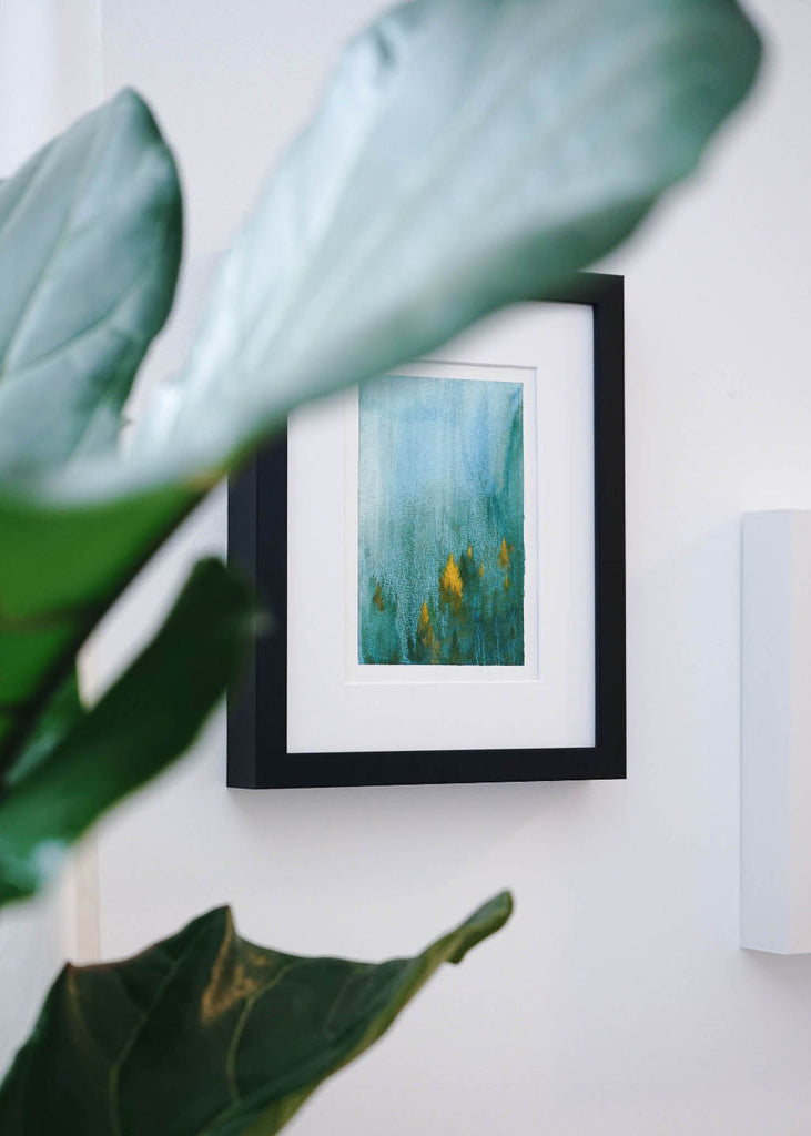 A beautiful green misty forest painting framed in a black IKEA RIBBA frame hanging on the white wall.